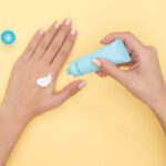 Close-up of a woman's hands applying cream on a pastel yellow background. Ideal for beauty and skincare themes.